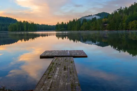 A wooden dock extends into a calm lake at sunset, surrounded by forest and mountains.