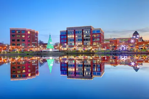 Colorful buildings and a Christmas tree reflected in calm water at dusk.