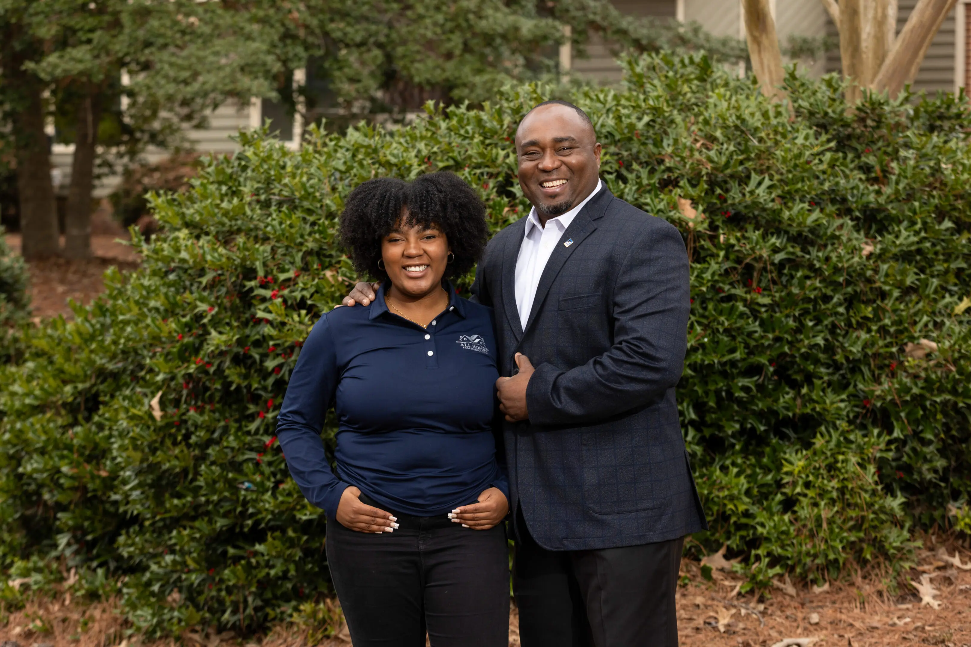A smiling couple standing outdoors with greenery in the background.