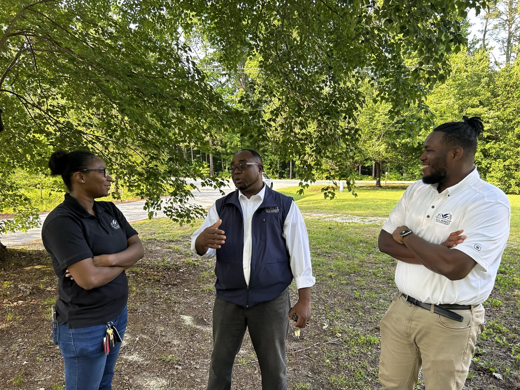 Three men engaged in a conversation outdoors under a large tree.