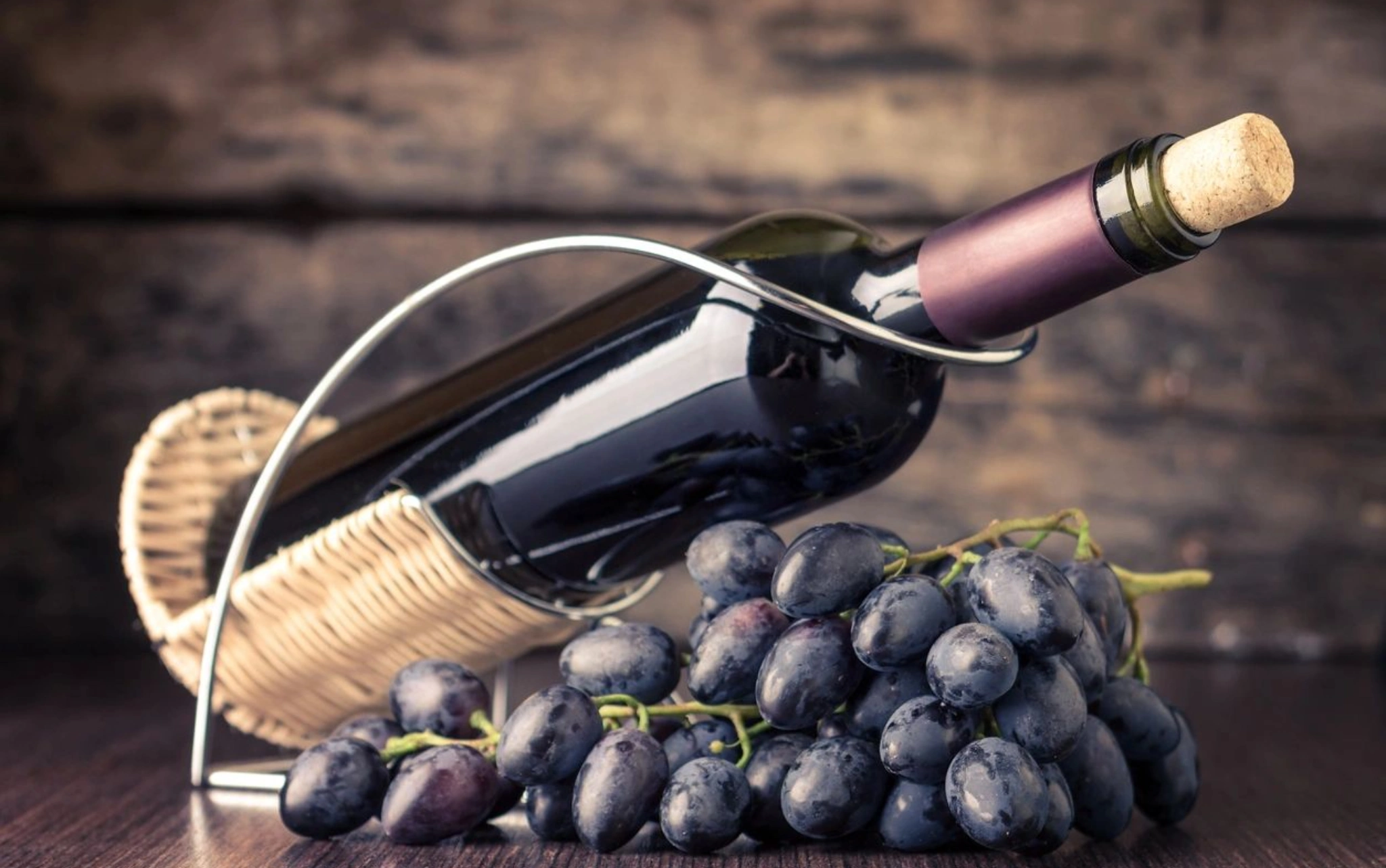 A bottle of red wine with grapes and corks on a rustic surface.